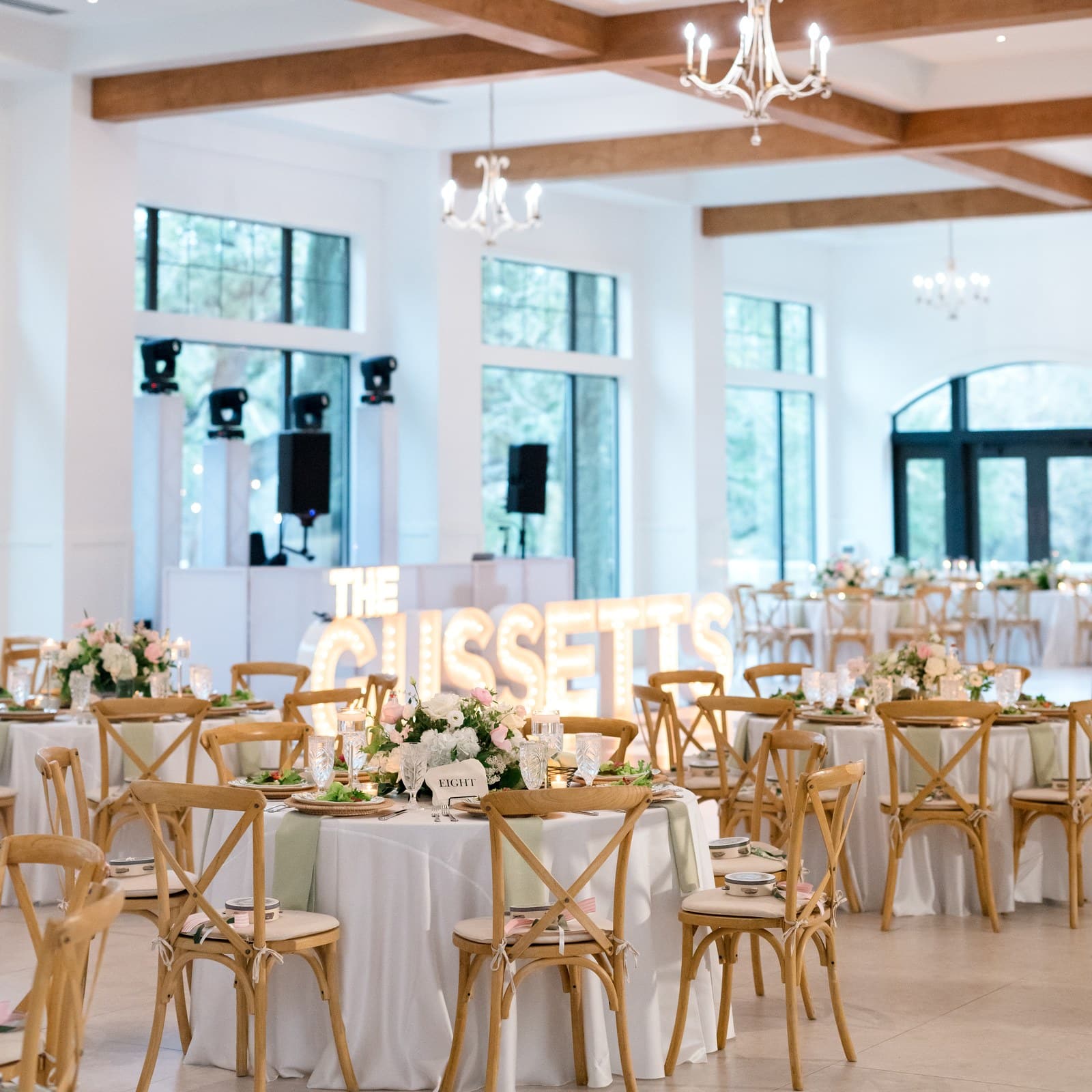 Ballroom reception tables with white linens beneath wood beams and chandeliers, with lighted marquee letters by the DJ stage