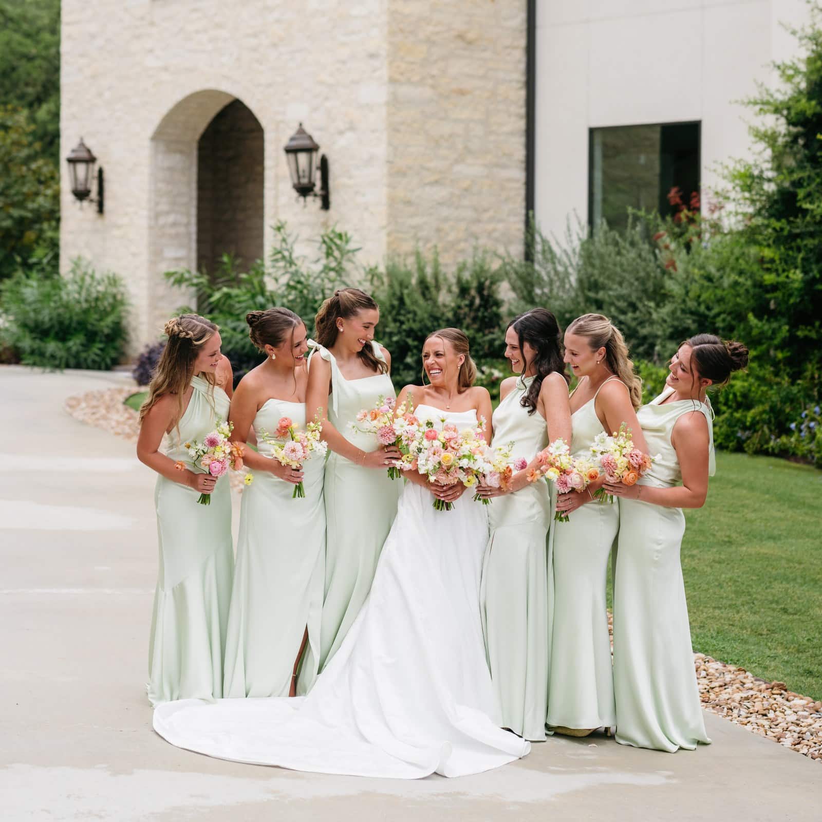 Bride and bridesmaids in sage green dresses laughing outside the stone entrance of The Preserve at Canyon Lake